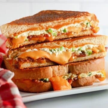 Stacked Jalapeno Popper Grilled Cheese sandwiches with melted cheese and green peppers on a white plate, next to a red cloth.