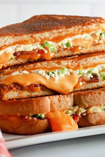 Stacked Jalapeno Popper Grilled Cheese sandwiches with melted cheese and green peppers on a white plate, next to a red cloth.
