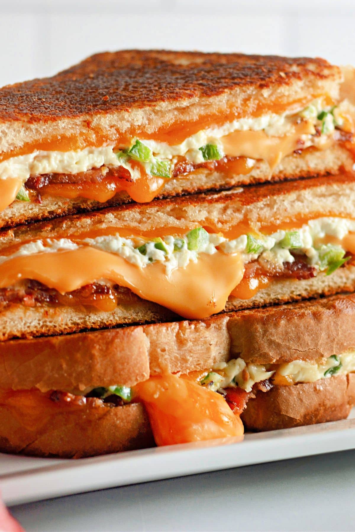 Close-up of stacked grilled cheese sandwiches with melted cheese, jalapeños, and crispy bread on a white plate.