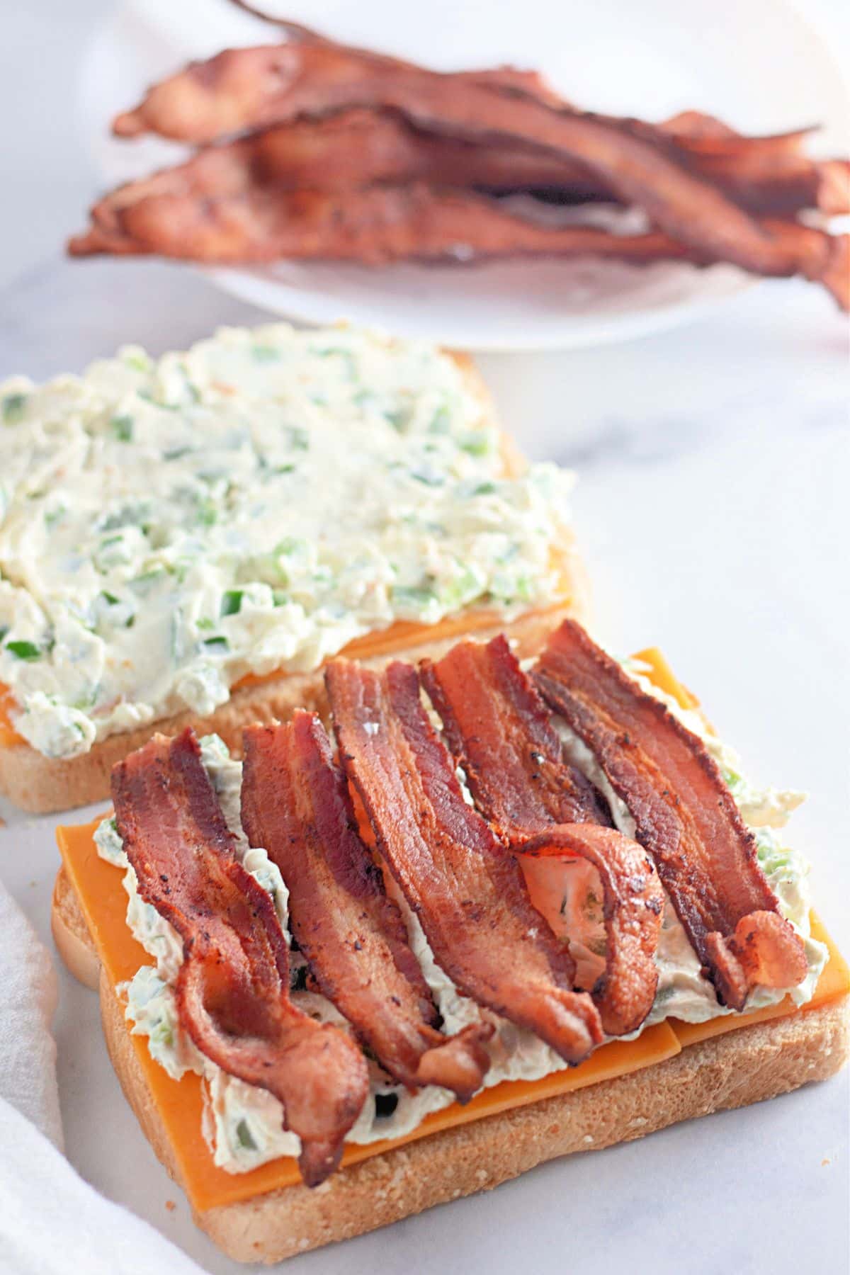 Open-faced sandwich with bacon, cheddar cheese, and creamy spread on white bread; more bacon in background.