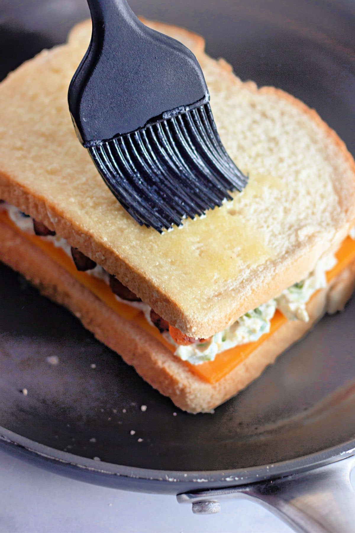 A pastry brush spreads butter on a sandwich in a frying pan, ready to be grilled.