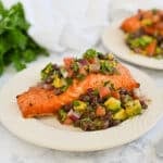 Grilled salmon fillet topped with avocado, black bean salsa on a white plate, parsley in the background.