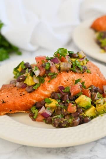 Grilled salmon fillet topped with avocado, black bean salsa on a white plate, parsley in the background.