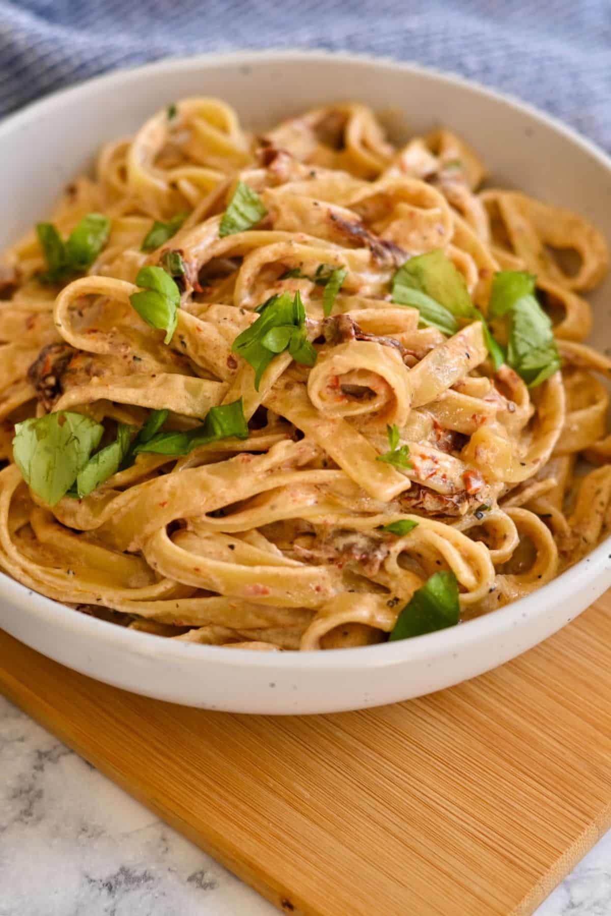 Creamy fettuccine pasta with herbs and sun-dried tomatoes, served in a white bowl on a wooden board.