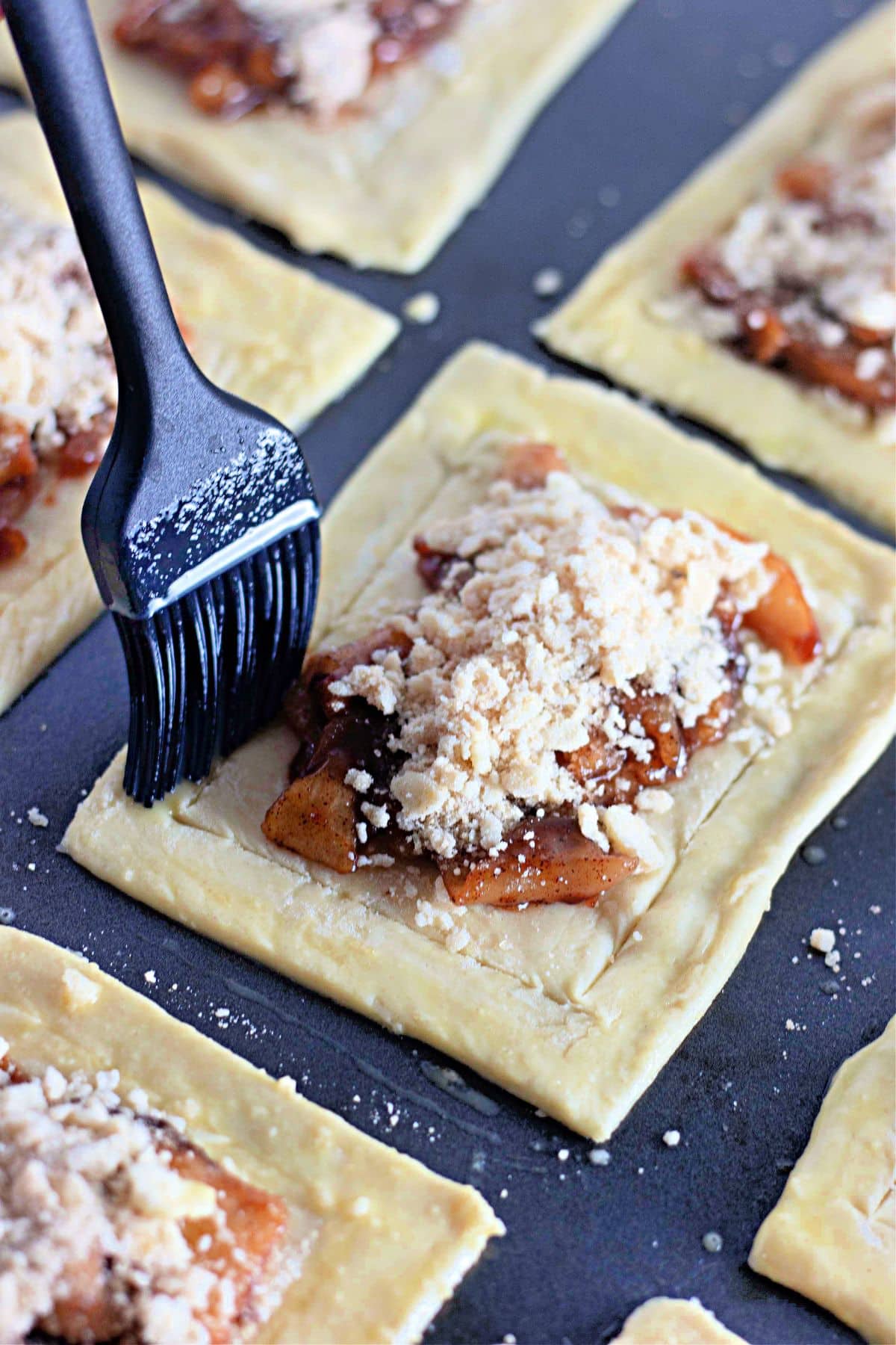 A pastry brush applies egg wash to puff pastry topped with apple filling and crumble on a baking sheet.