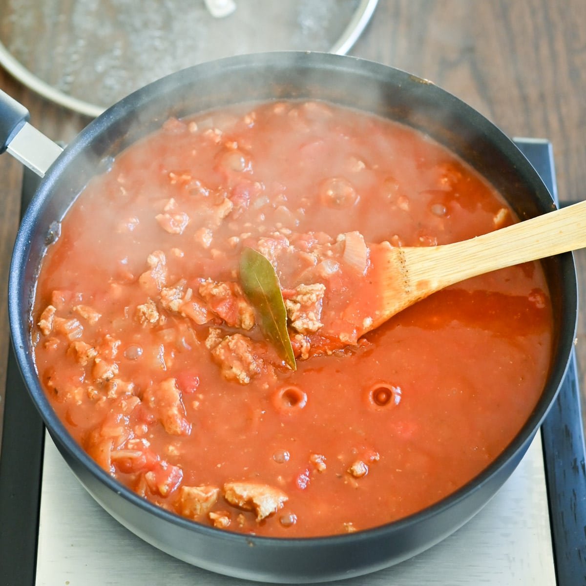 A pot of simmering tomato meat sauce with a bay leaf, being stirred with a wooden spoon.