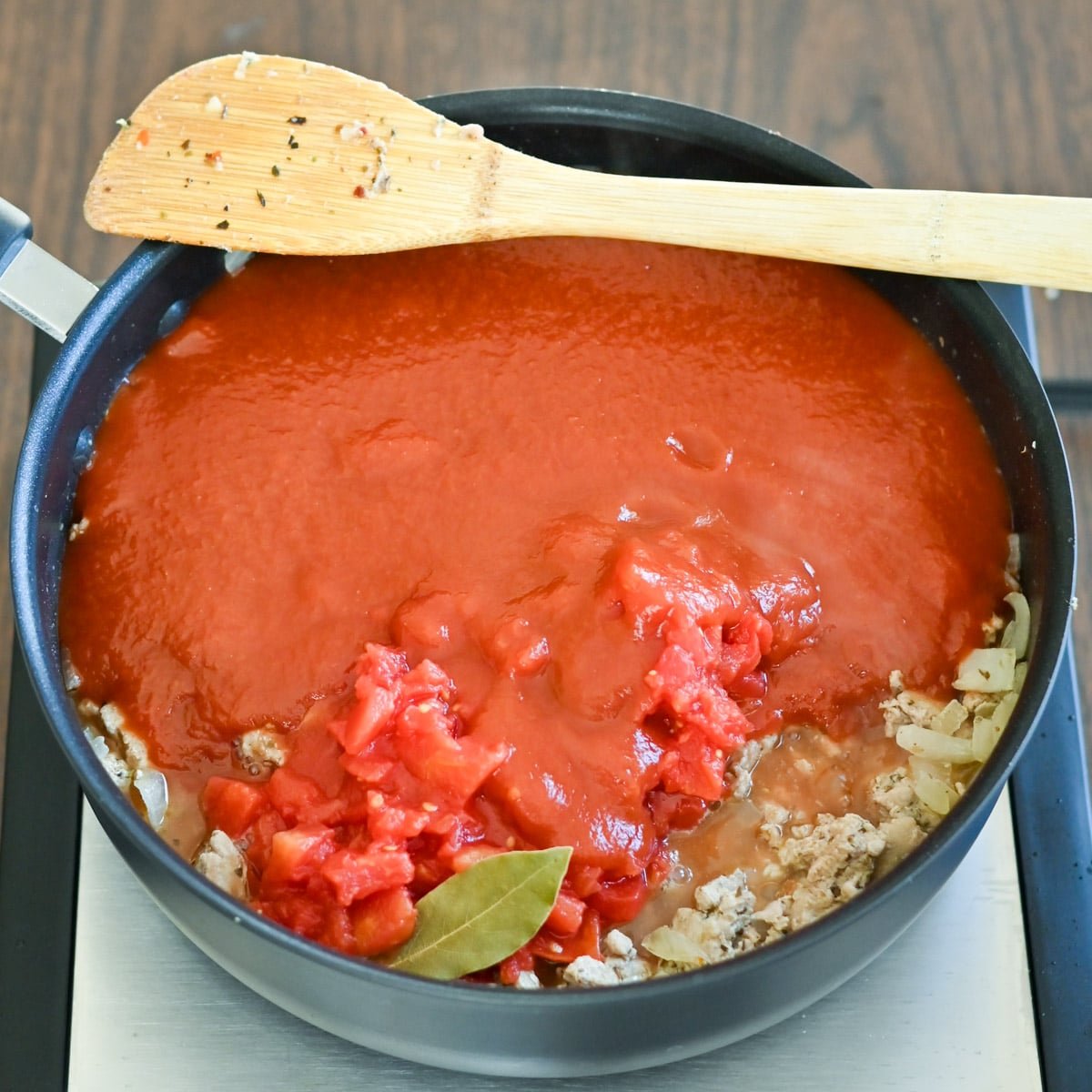 Pot of tomato sauce with diced tomatoes, ground meat, onions, bay leaf, and a wooden spoon on top.