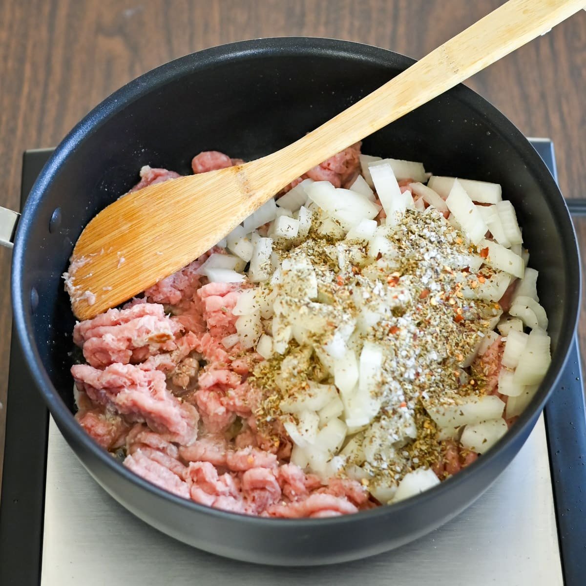 Ground meat, chopped onions, and spices in a black pot being stirred with a wooden spoon on a stovetop.