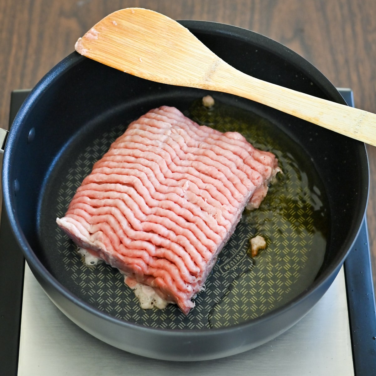 Raw ground meat in a skillet with oil, being cooked with a wooden spatula on a stovetop.