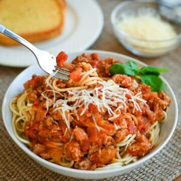 A bowl of spaghetti with meat sauce, topped with grated cheese, with garlic bread and cheese in the background.