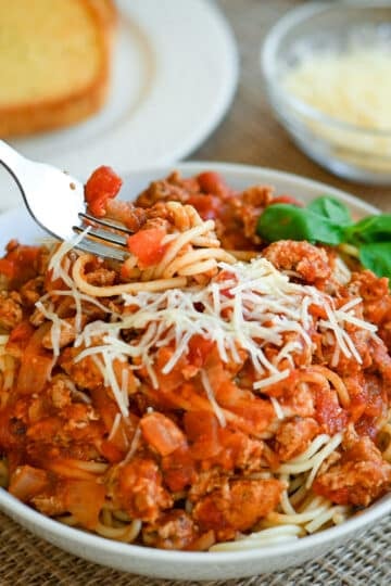 A bowl of spaghetti with meat sauce, topped with grated cheese, with garlic bread and cheese in the background.