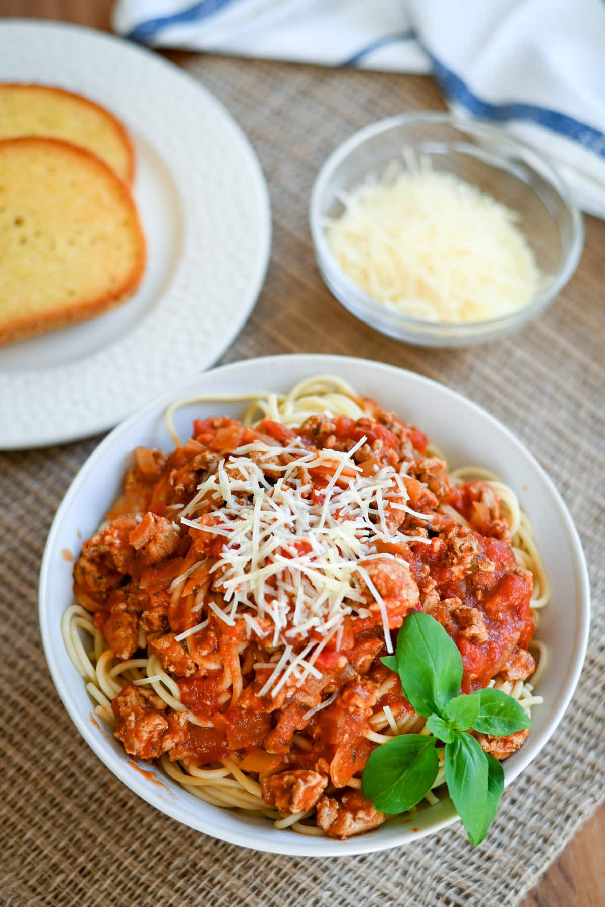 A bowl of spaghetti with meat sauce and cheese, garnished with basil, beside garlic bread and grated cheese.