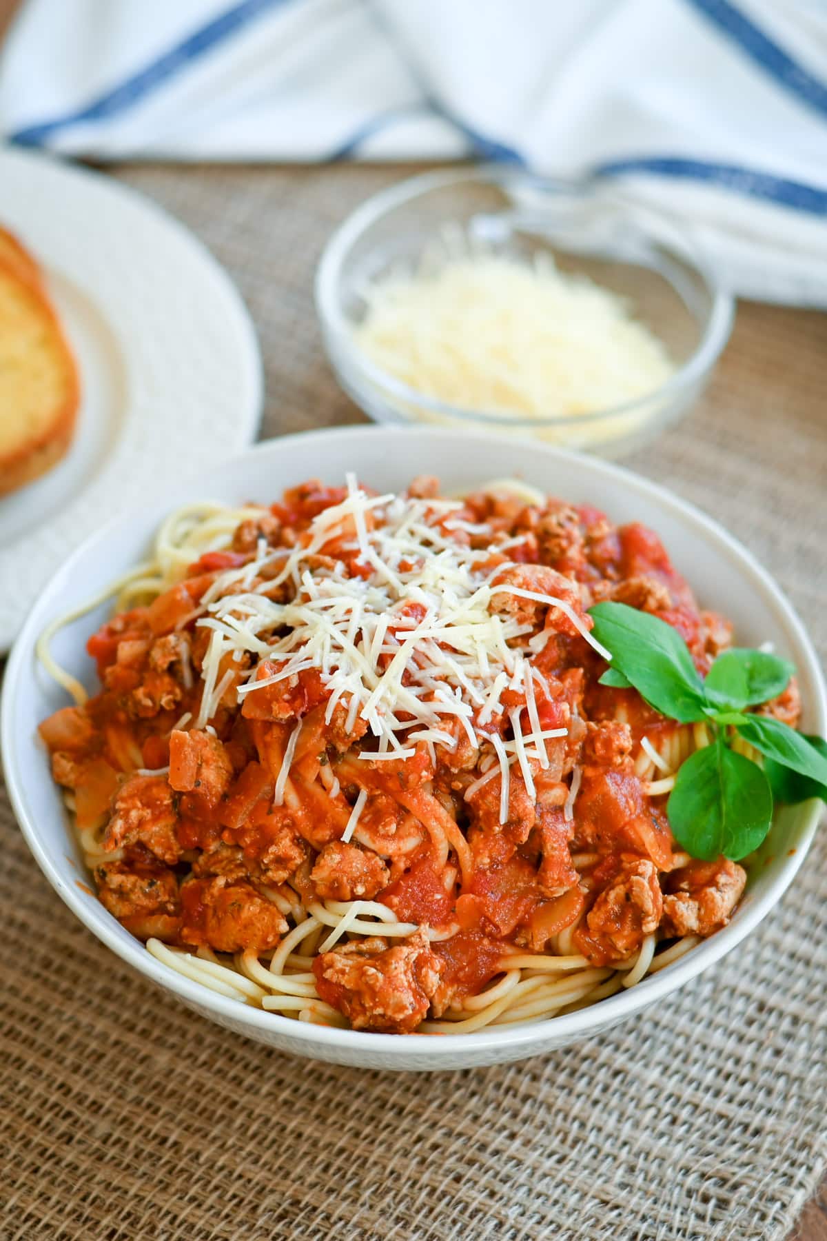 Bowl of spaghetti with meat sauce, topped with grated cheese and basil, with garlic bread and cheese in background.
