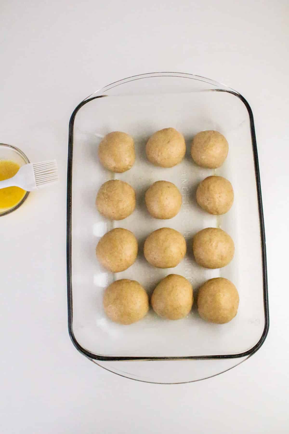 A glass dish with 12 dough balls and a bowl of egg wash with a brush on a white surface.