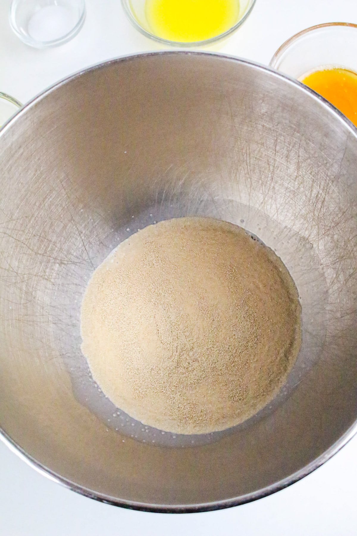 A metal mixing bowl with yeast and sugar inside, surrounded by bowls of melted butter and eggs.