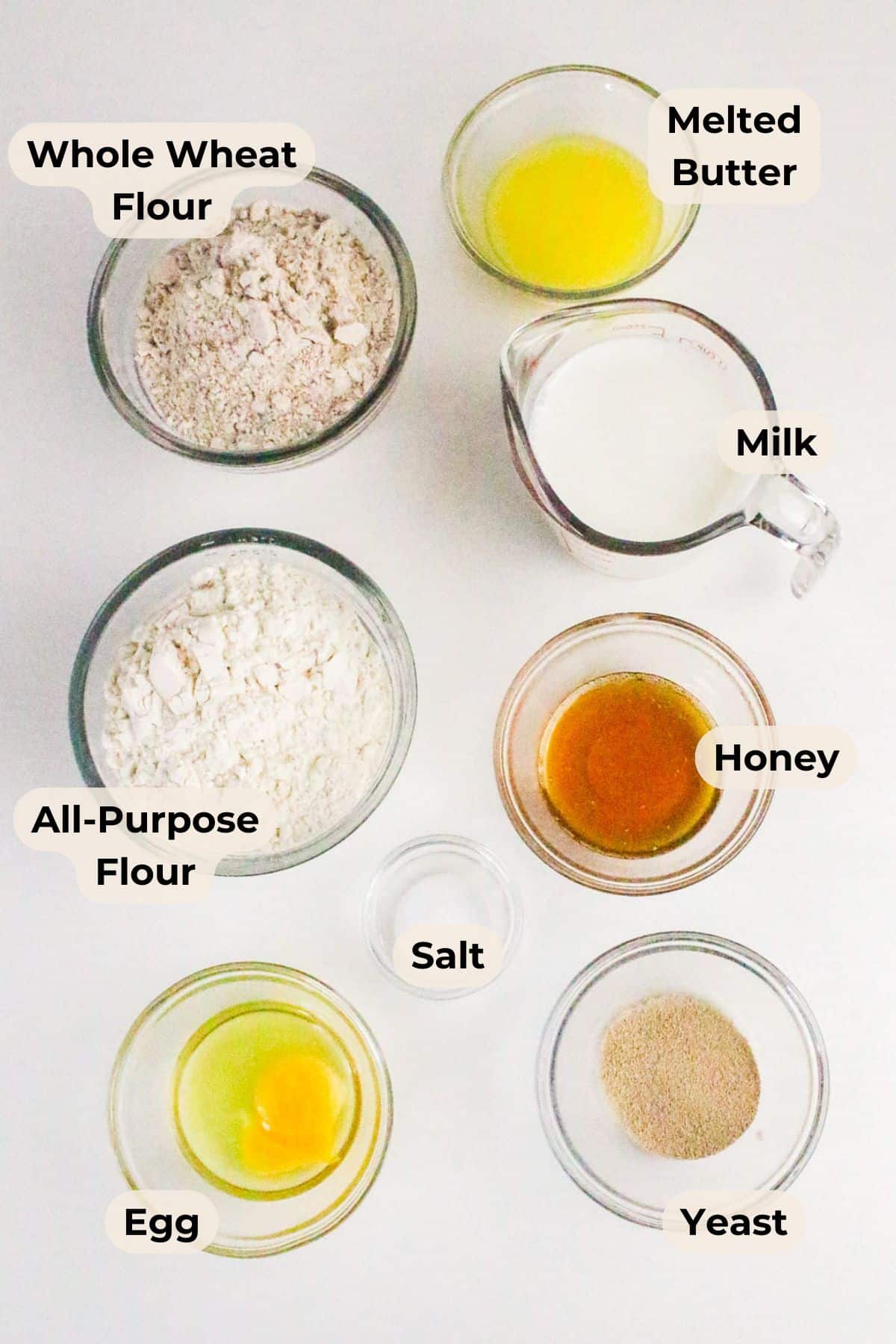 Top-down view of labeled bowls containing flour, butter, milk, honey, salt, yeast, and an egg on a white surface.