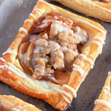 A rectangular apple pastry with crumble topping and white icing drizzle on a metal baking sheet.