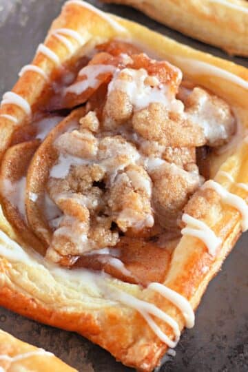 A rectangular apple pastry with crumble topping and white icing drizzle on a metal baking sheet.