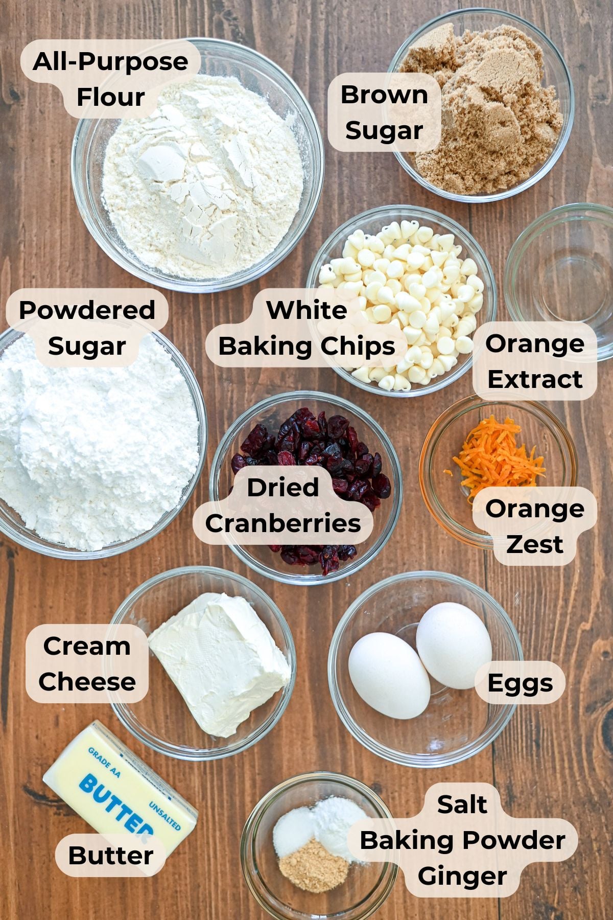 Top-down view of labeled bowls containing cookie ingredients, including flour, sugars, chips, eggs, butter, and flavorings.