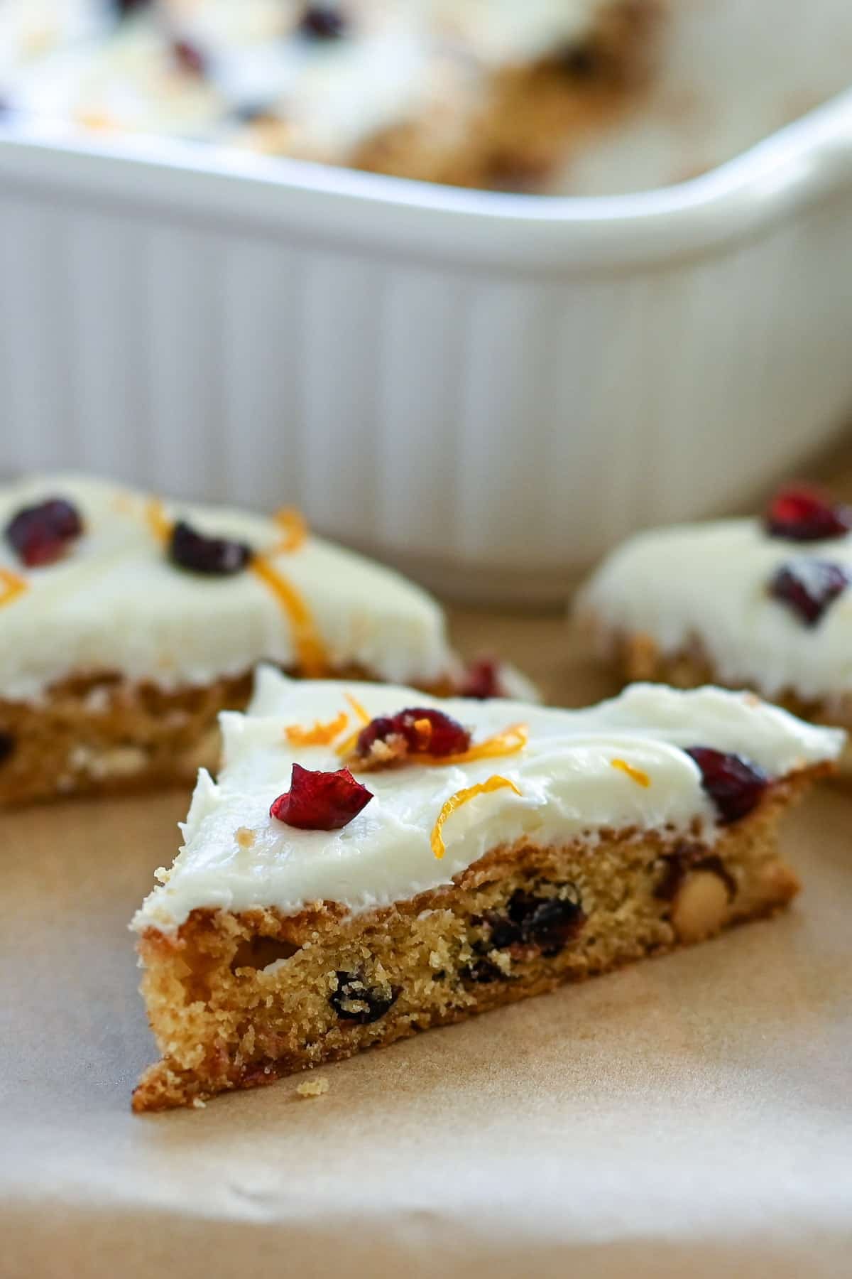 A cranberry bar topped with white icing, orange zest, and dried cranberries, with more bars in the background.