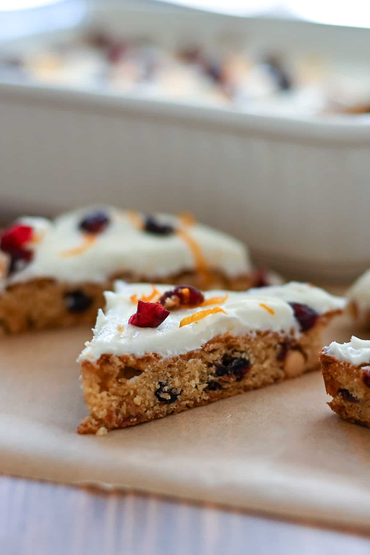 A frosted dessert bar with cranberries and orange zest, sliced and placed on parchment paper.