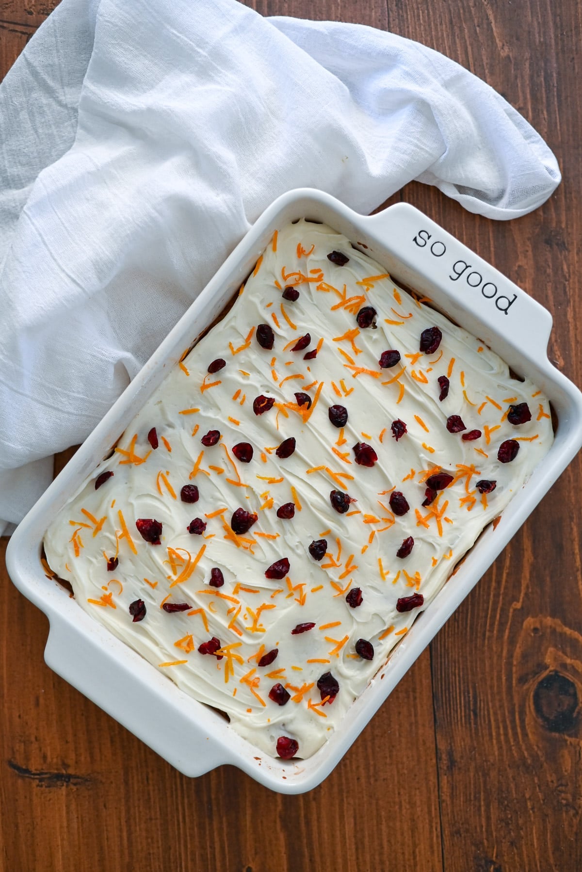 A frosted bar cookie topped with cranberries and orange zest in a white dish labeled "so good" on a wooden table.