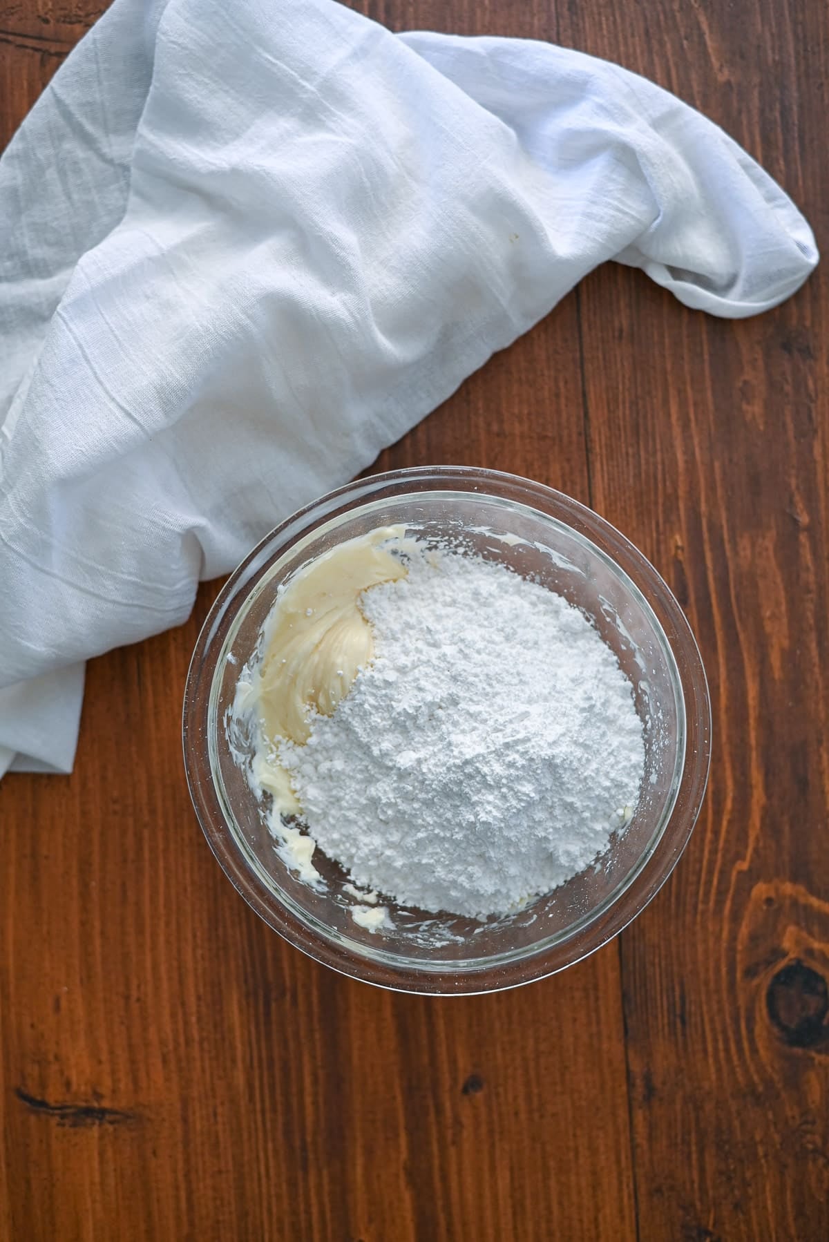 A glass bowl with powdered sugar and butter, next to a white cloth on a wooden surface.