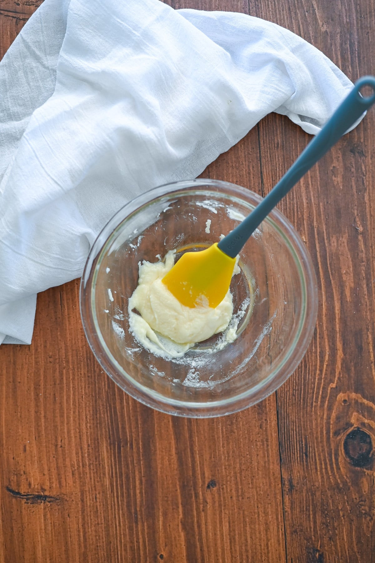 A glass bowl with a yellow spatula mixing batter, on a wooden table with a white cloth nearby.