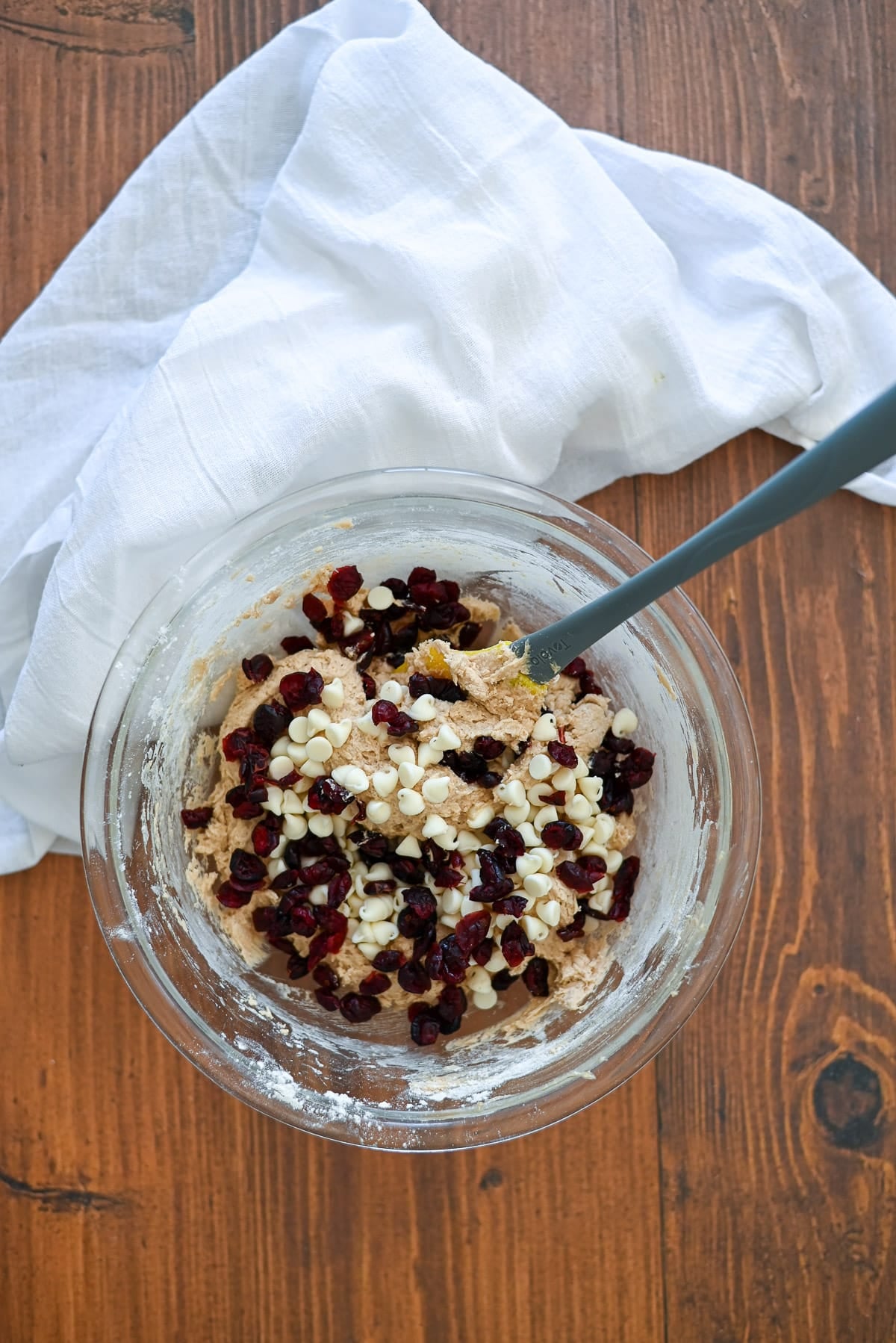 A glass bowl of cookie dough with white chocolate chips and dried cranberries, on a wooden table with a white cloth.