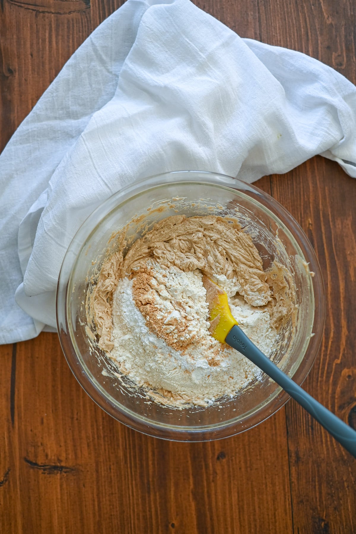 A mixing bowl with batter and flour, a spatula, and a white cloth on a wooden table.
