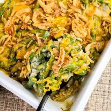 Green bean casserole topped with crispy fried onions and melted cheese in a white baking dish with a serving spoon.