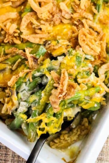 Green bean casserole topped with crispy fried onions and melted cheese in a white baking dish with a serving spoon.