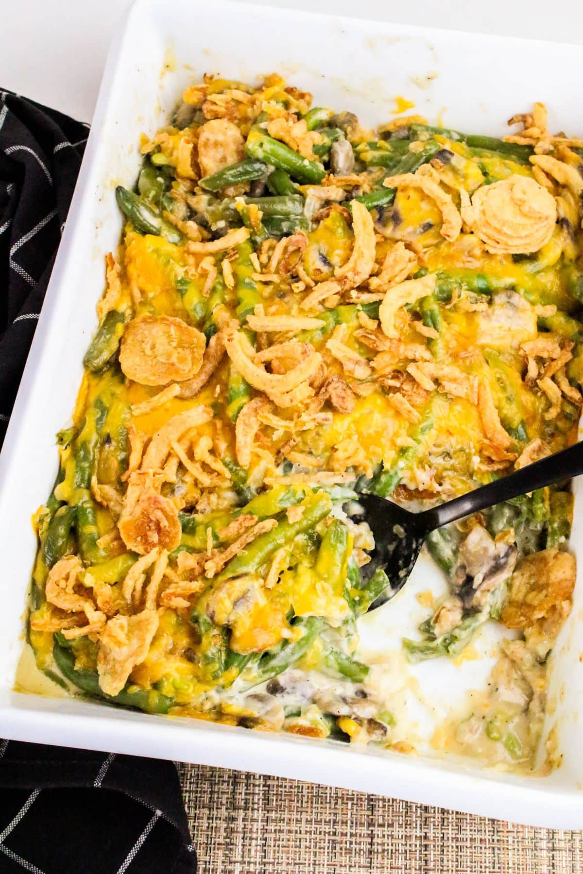 Green bean casserole topped with crispy fried onions in a white baking dish, partially served with a black spoon.