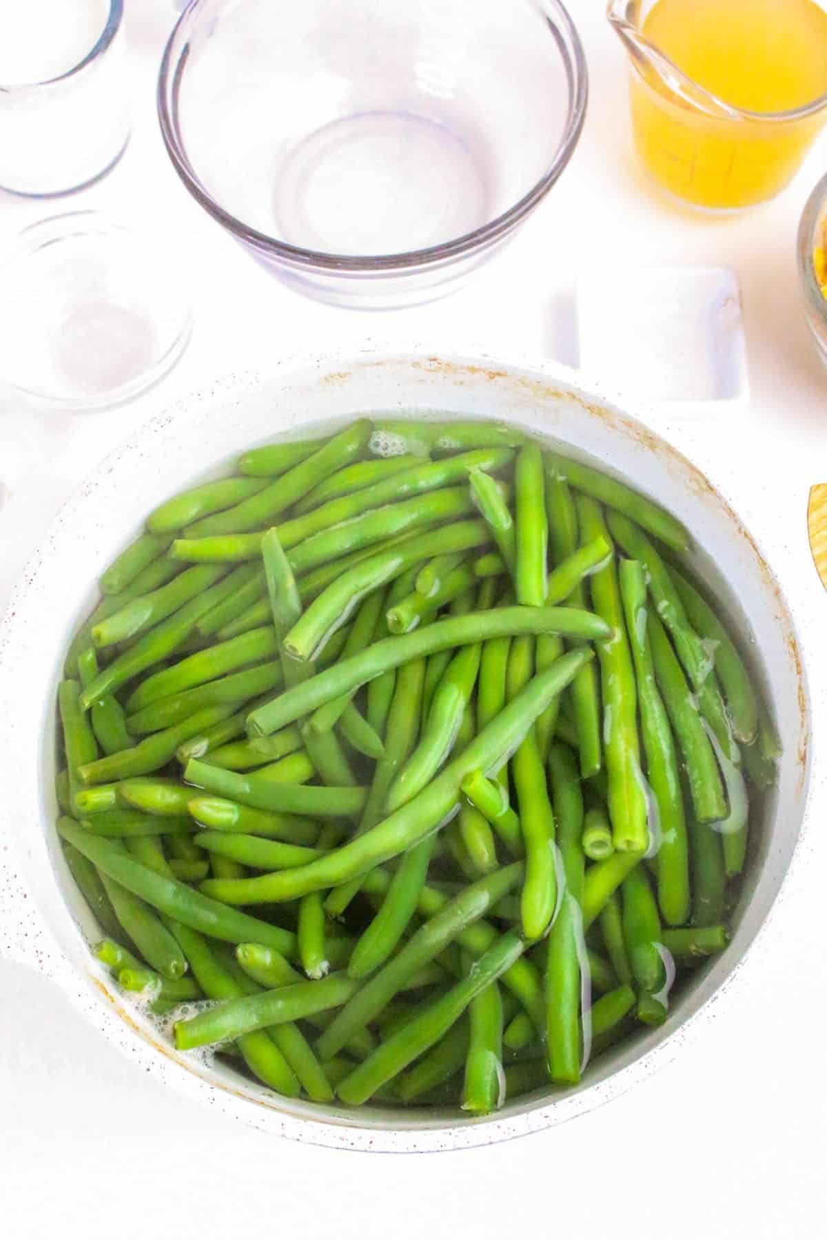 A pot filled with fresh green beans submerged in water, surrounded by empty glass bowls.