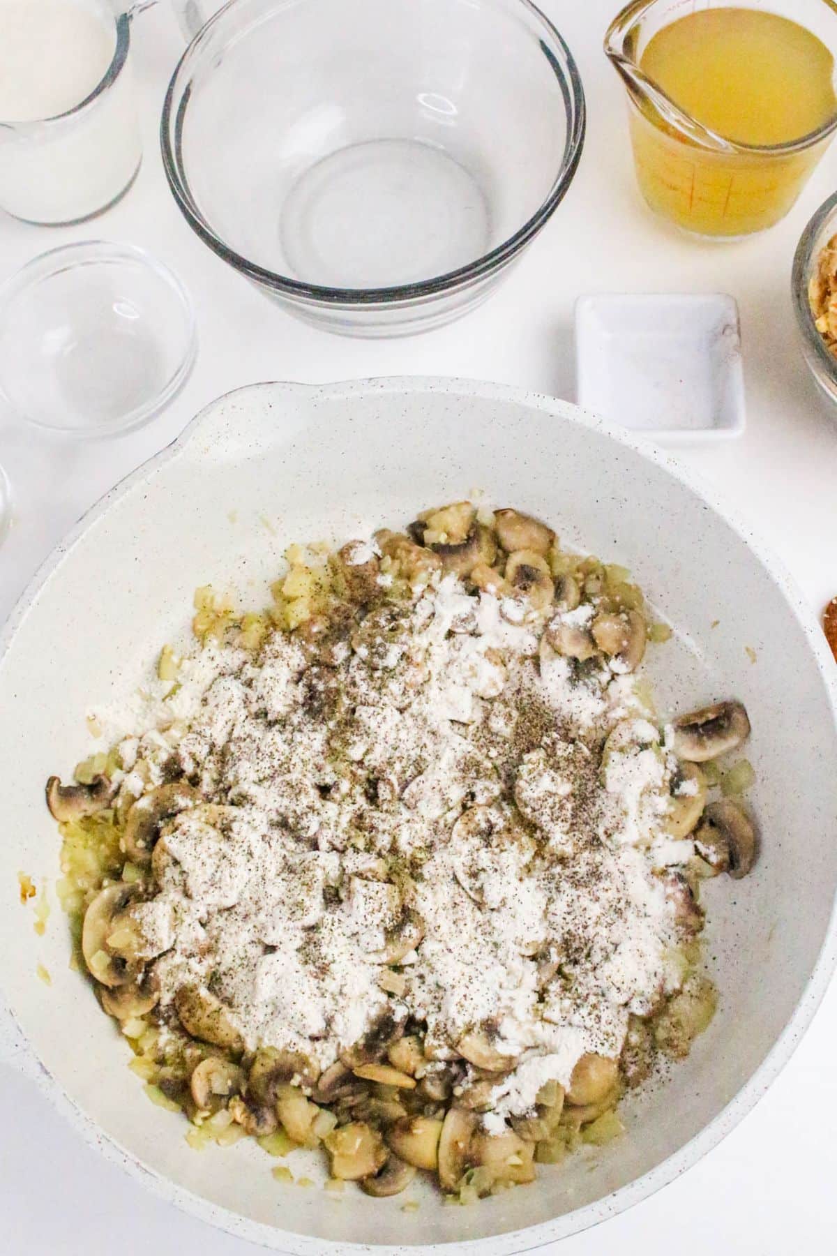 A white pan with mushrooms, onions, flour, and pepper, surrounded by bowls of ingredients on a white surface.