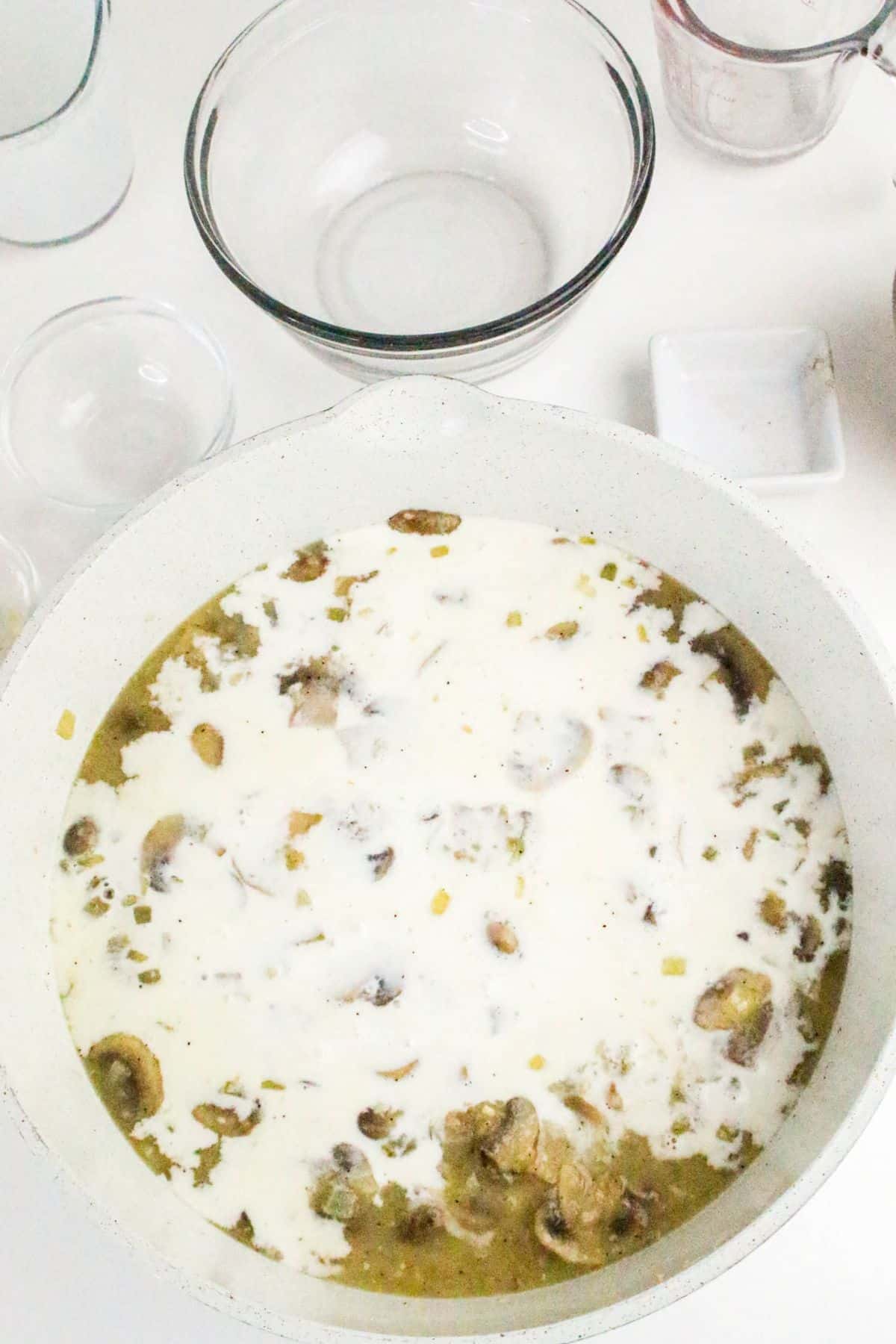 A white pot filled with creamy mushroom soup sits on a white surface near empty glass bowls.