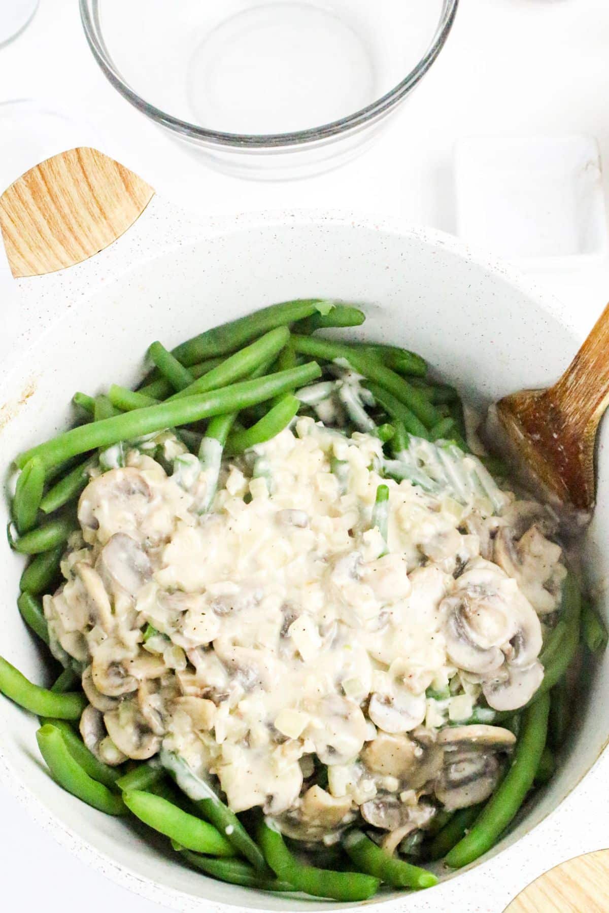 Green beans in a white pot topped with creamy mushroom sauce, stirred with a wooden spoon.