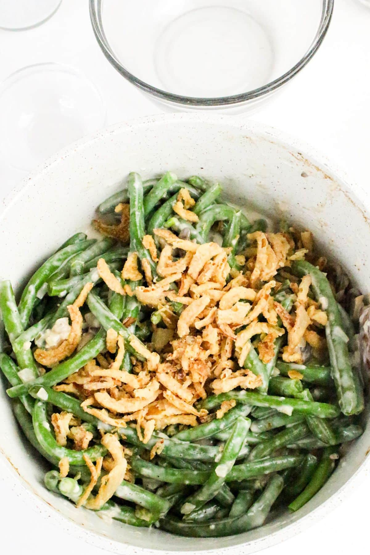 Green bean casserole topped with crispy fried onions in a white dish, with empty glass bowls nearby.