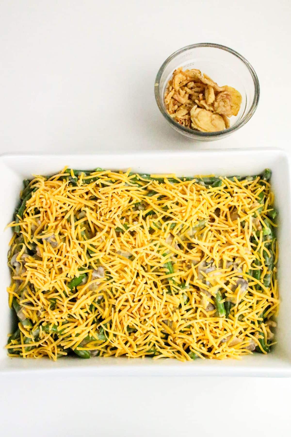 Rectangular dish with green bean casserole topped with shredded cheese, next to a bowl of crispy fried onions.