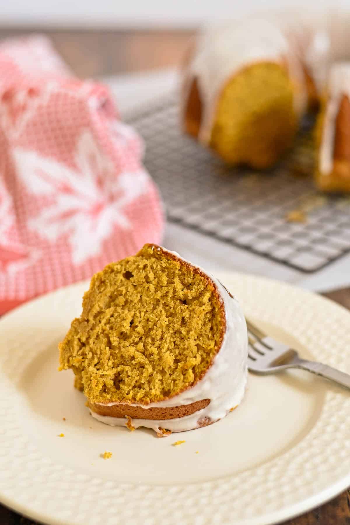 A slice of Bundt cake with white icing on a plate, with a fork and the rest of the cake in the background.