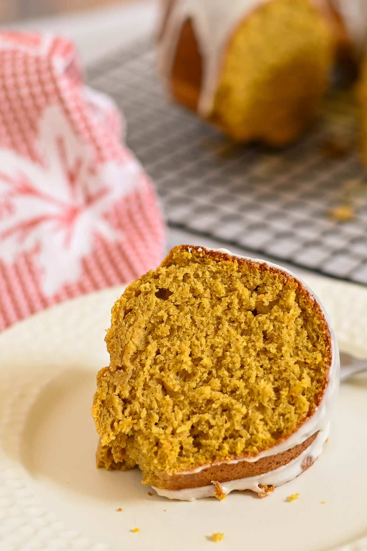 A slice of pumpkin bundt cake with white icing sits on a plate, with a red towel in the background.