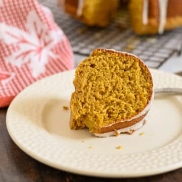 A slice of pumpkin bundt cake with icing on a white plate, a red towel in the background.