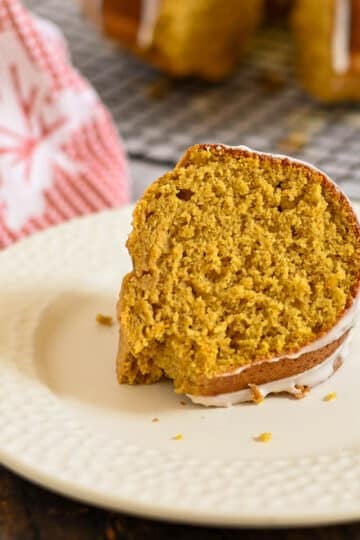 A slice of pumpkin bundt cake with icing on a white plate, a red towel in the background.