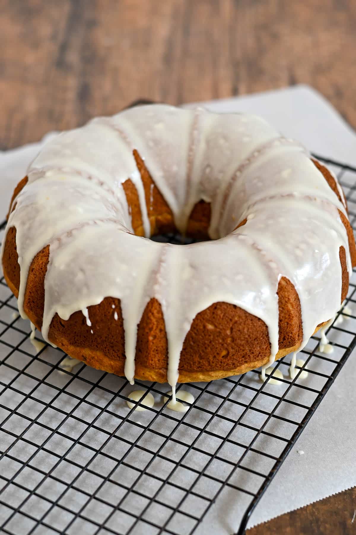A bundt cake with white glaze icing sits on a cooling rack over parchment paper.