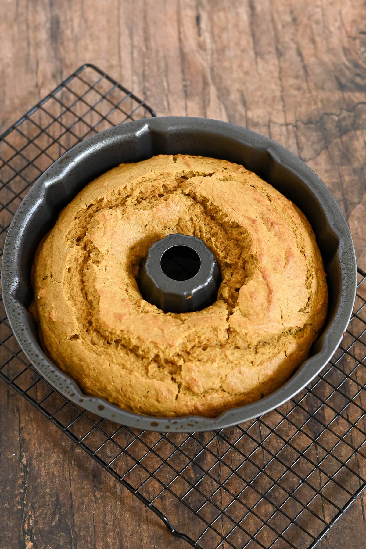 A Bundt cake in a pan cooling on a wire rack atop a wooden surface.