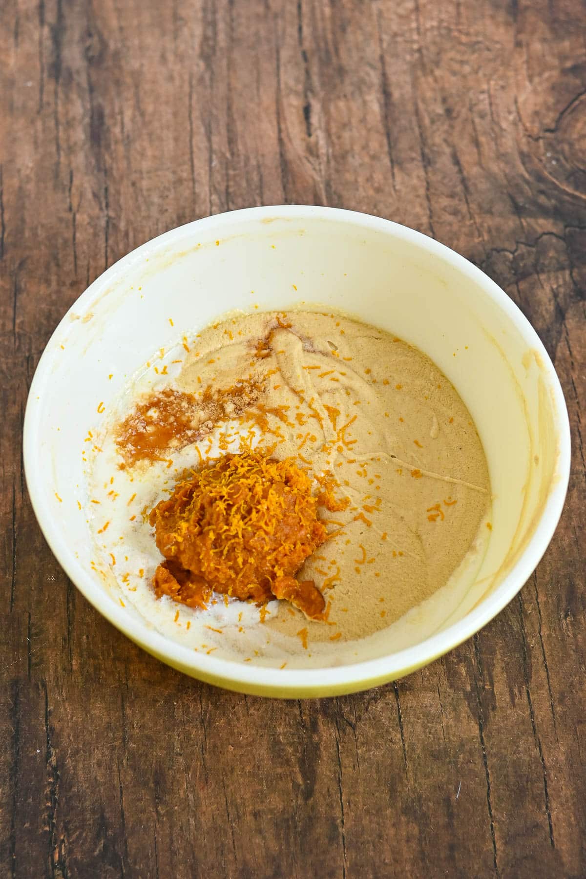 A white bowl with brown sugar, spices, and orange zest on a wooden surface.