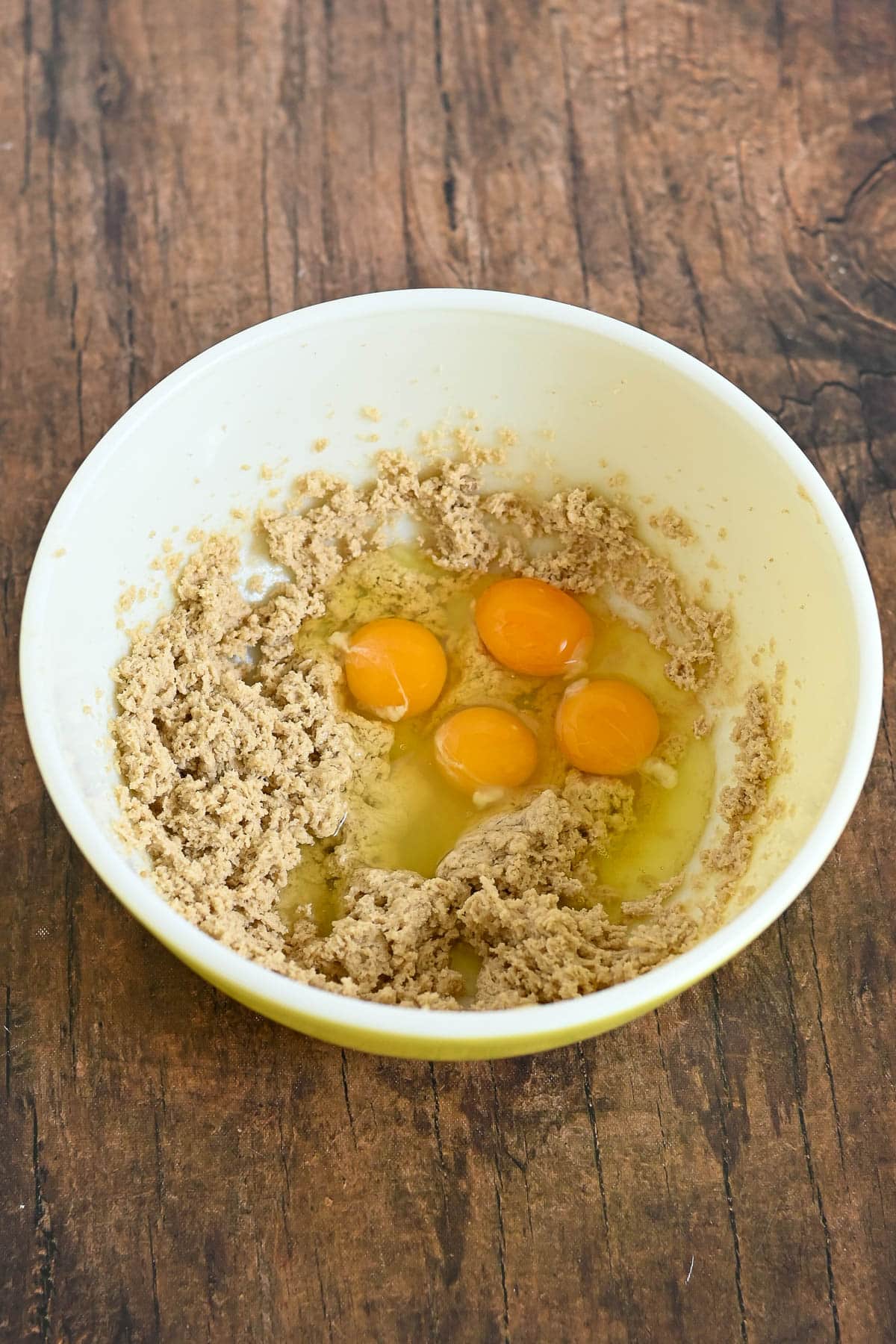 A mixing bowl with creamed sugar and butter, and four cracked eggs on top, on a wooden surface.