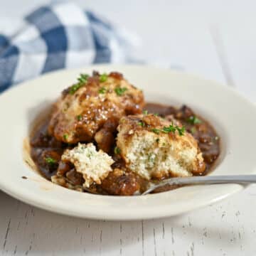 A bowl of stew topped with two dumplings, garnished with herbs, with a spoon and a plaid napkin nearby.