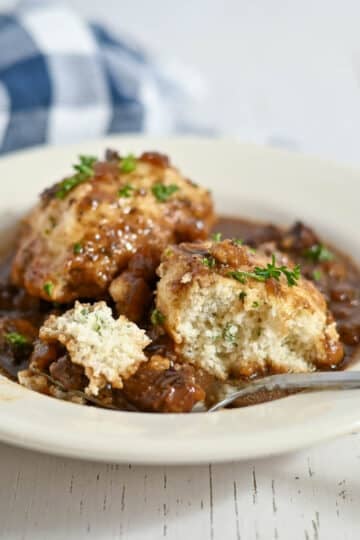 A bowl of stew topped with two dumplings, garnished with herbs, with a spoon and a plaid napkin nearby.