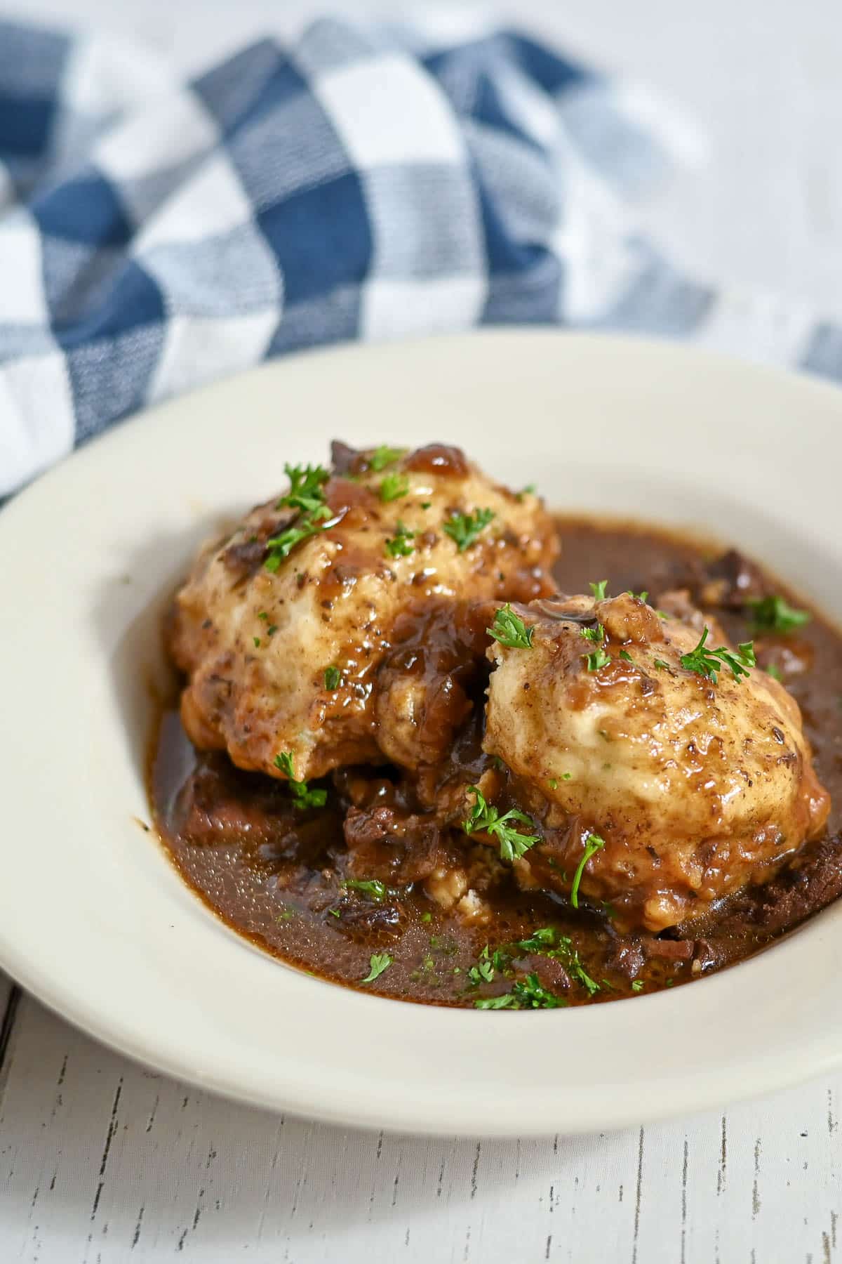 Two dumplings topped with gravy and herbs on a white plate, with a checkered cloth in the background.